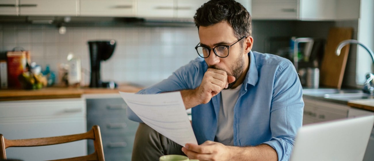 Man staring at paper electric bill with laptop open on kitchen table.